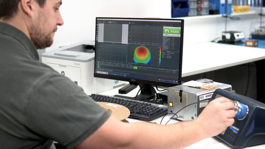 A technician in a laboratory or workshop setting operates fibre optic testing equipment connected to a computer. The monitor displays measurement graphs and a green “PASS” result, indicating the tested component meets the required standard.